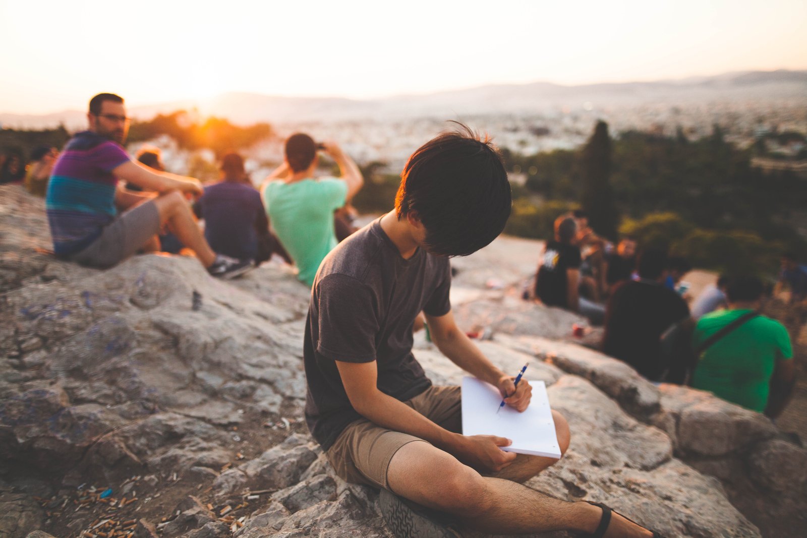 Teenage or young adult-aged boy writing or drawing in a journal or sketch book alone while a group sits nearby