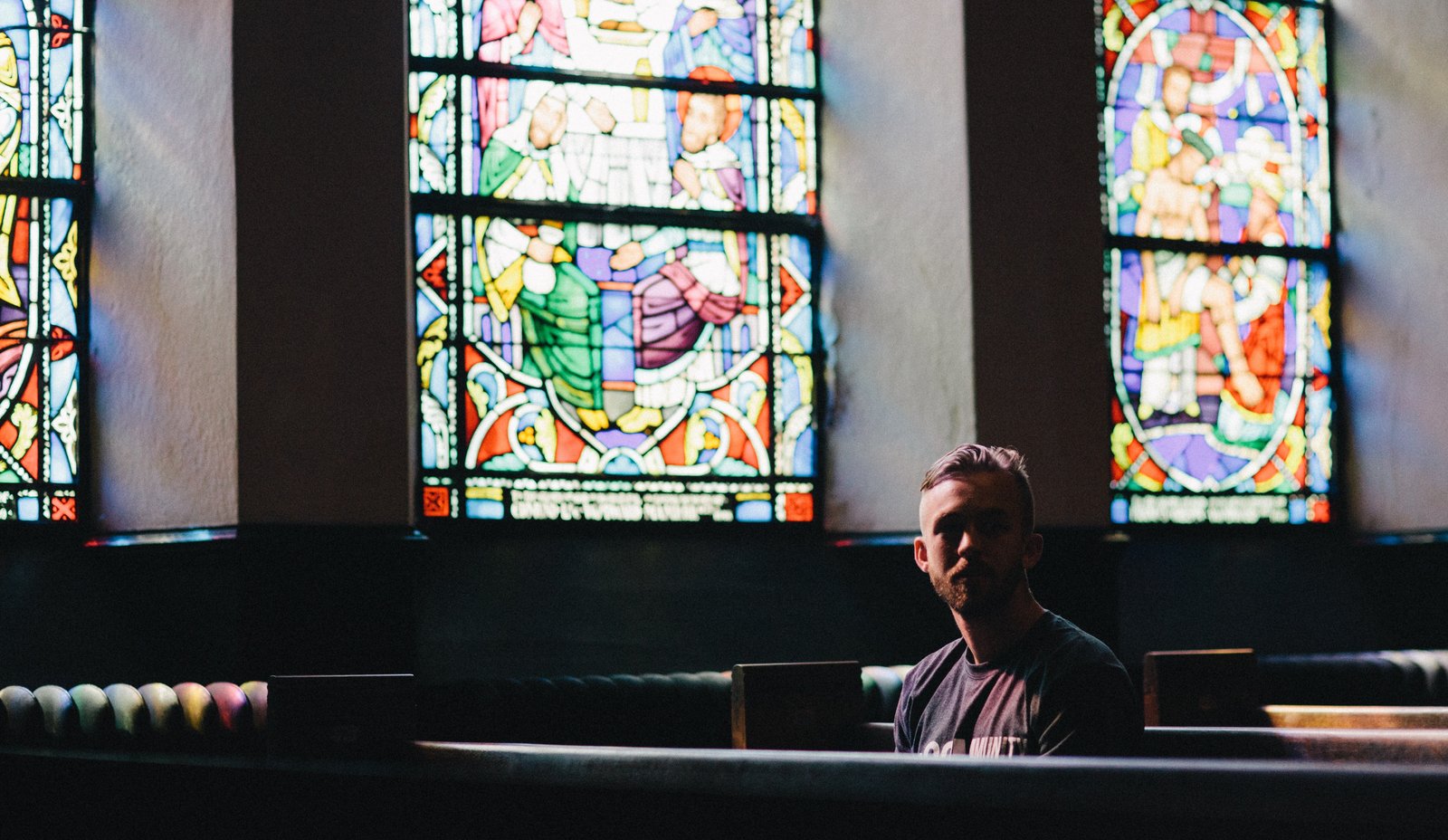 White male with beard, sitting in church pew alone, surrounded by stained glass windows
