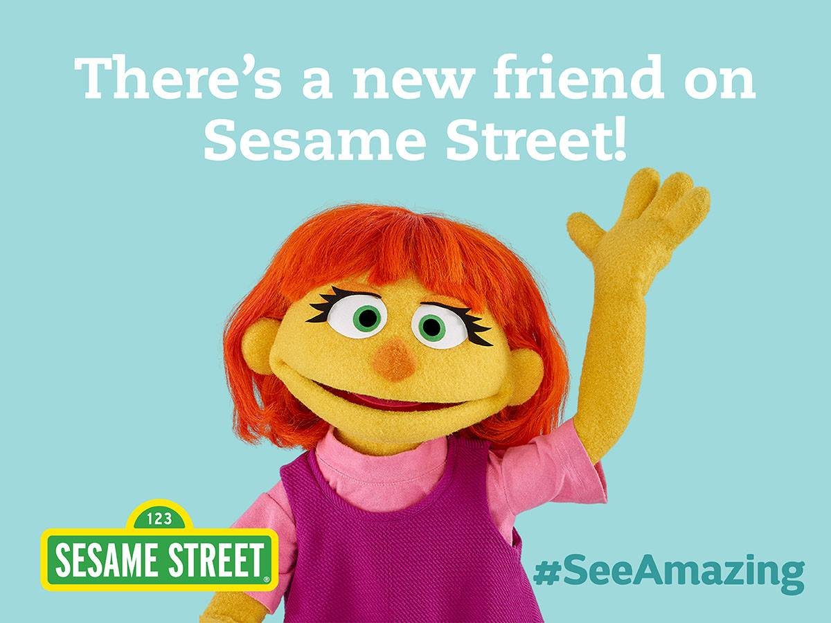 A marketing image from Sesame Street of Julia, a new muppet on the autism spectrum. She is wearing a pink shirt and jumper, waving gesture with a smile. Above her are the words "There's a new friend on Sesame Street!" in white lettering. Bottom right, the words # see amazing in turquoise.