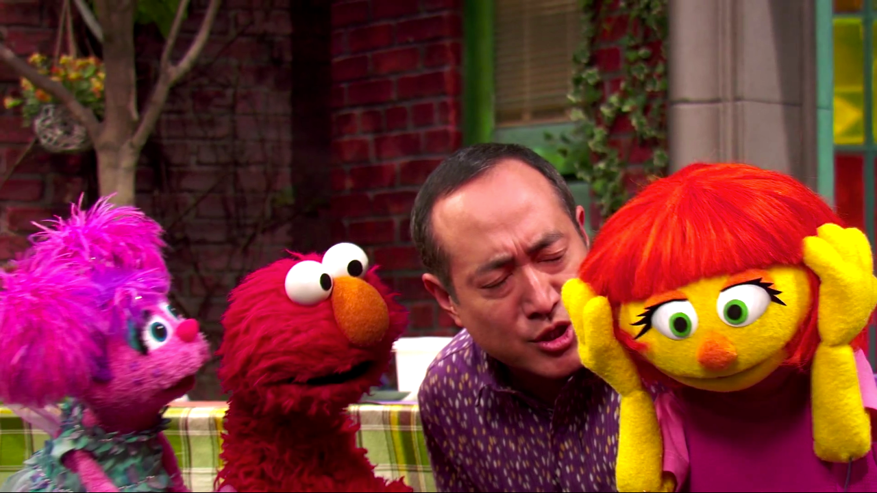 Scene from the new Sesame Street episode introducing Julia. Abby, Elmo, and a human friend look at Julia with concern, while she covers her ears with her hands.