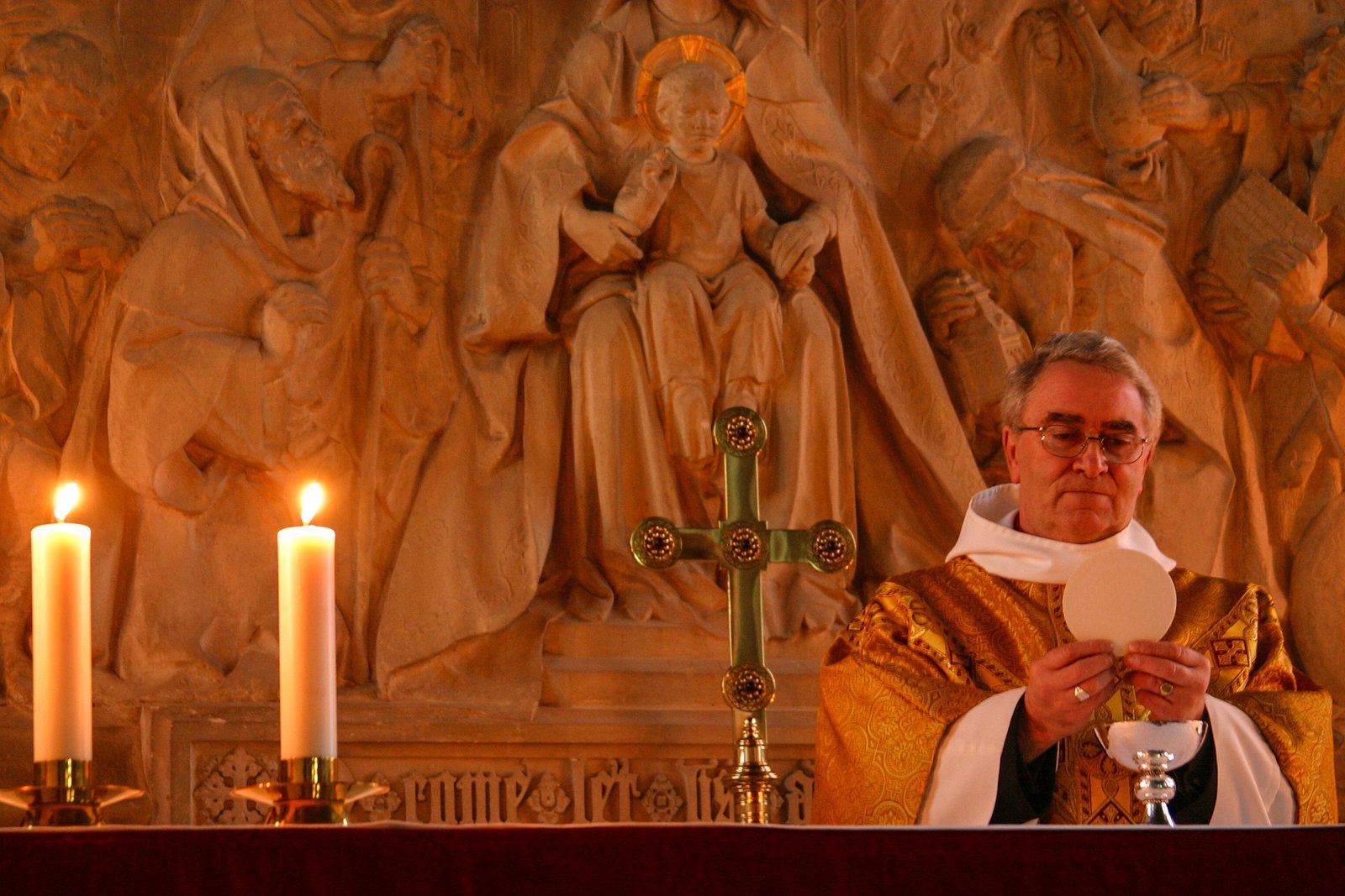 Priests in gold vestments holding Eucharist. Two glowing candles and simple gold cross on altar. Relief of child, Jesus on wall behind altar and priest.